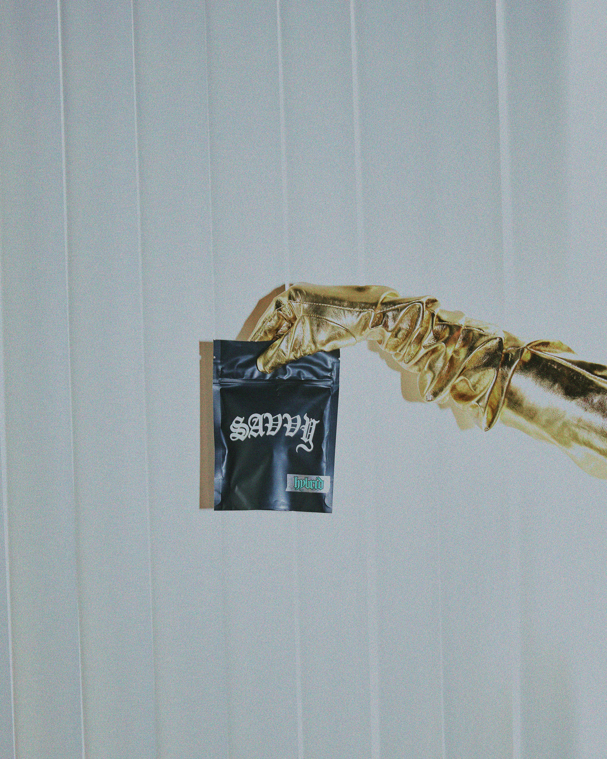 Shiny gold gloved hand holds a black sealed product bag with the word "Savvy" against a ridged white backdrop in a studio shot.