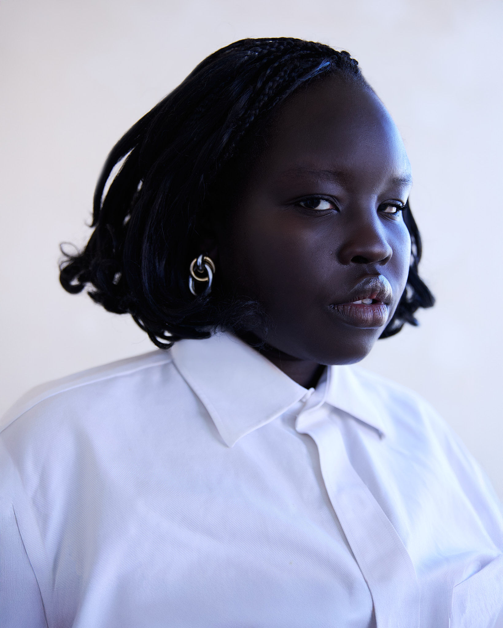 Portrait of an adult female with dark skin, wearing a white collared shirt, silver earrings, and with black braided hair against a plain wall.