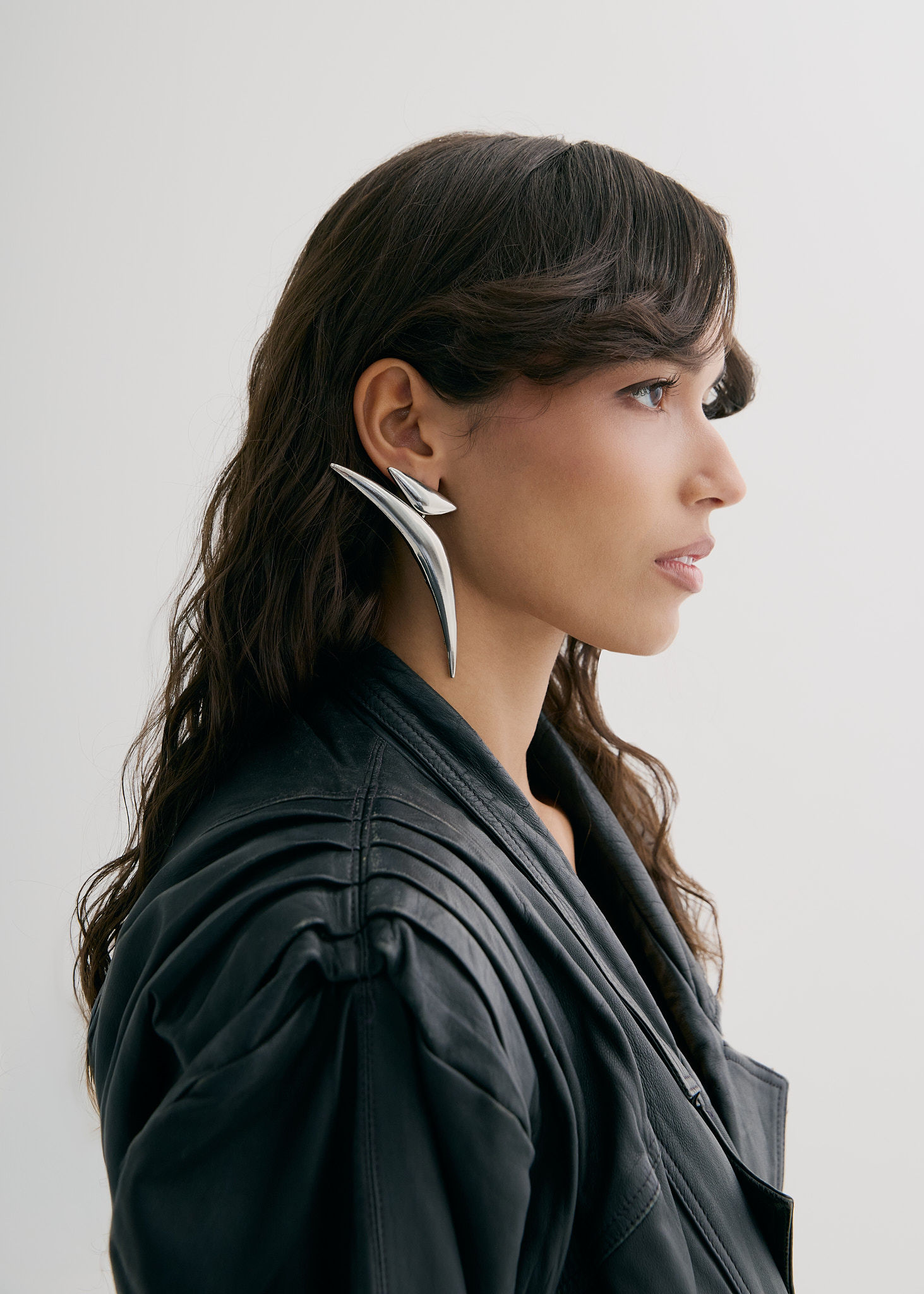 Profile shot of a woman with dark wavy hair, wearing a black jacket and silver sculptural earrings against a neutral background.