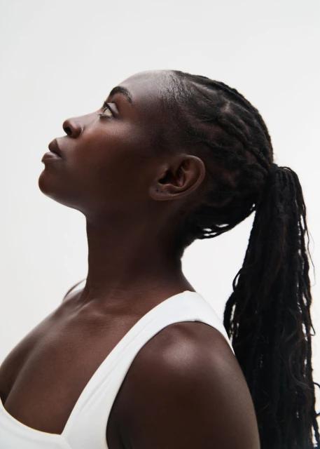 Profile of a young Black woman with braided hair, looking upwards against a plain white backdrop in a beauty or fashion context.