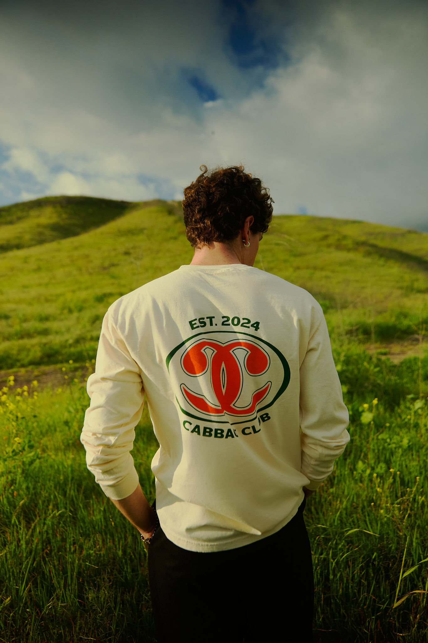 A person stands in a green field with a t-shirt displaying 'Cabba Club' and 'Est. 2024', against a backdrop of rolling hills and cloudy sky.