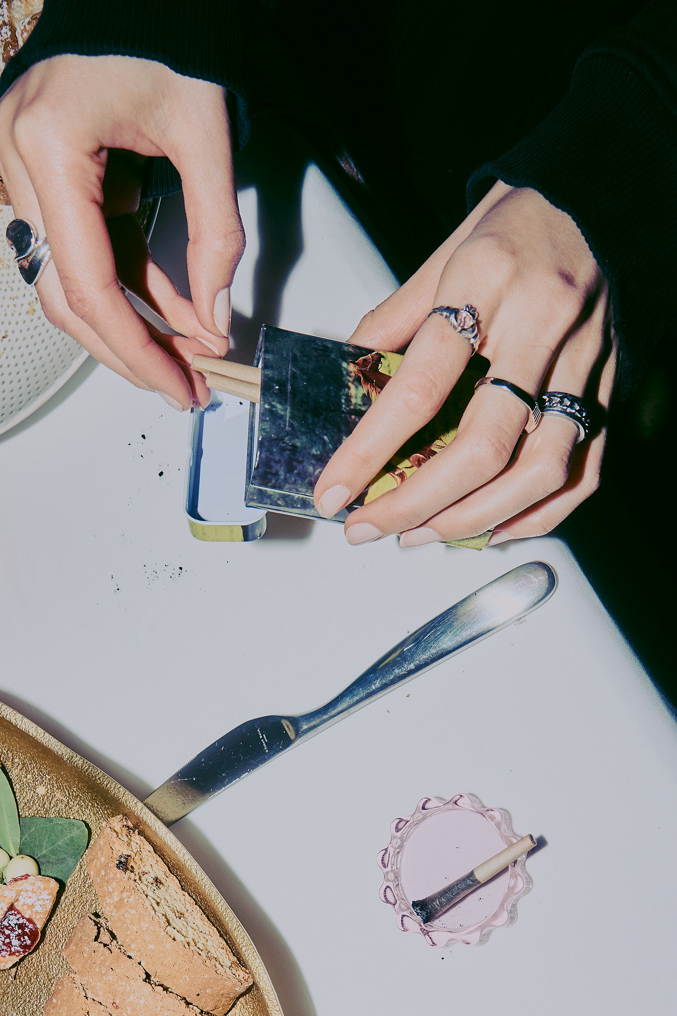 Overhead view of hands rolling cigarettes at a table with a tin, lighter, bread, and knife, showcasing a lifestyle setting.