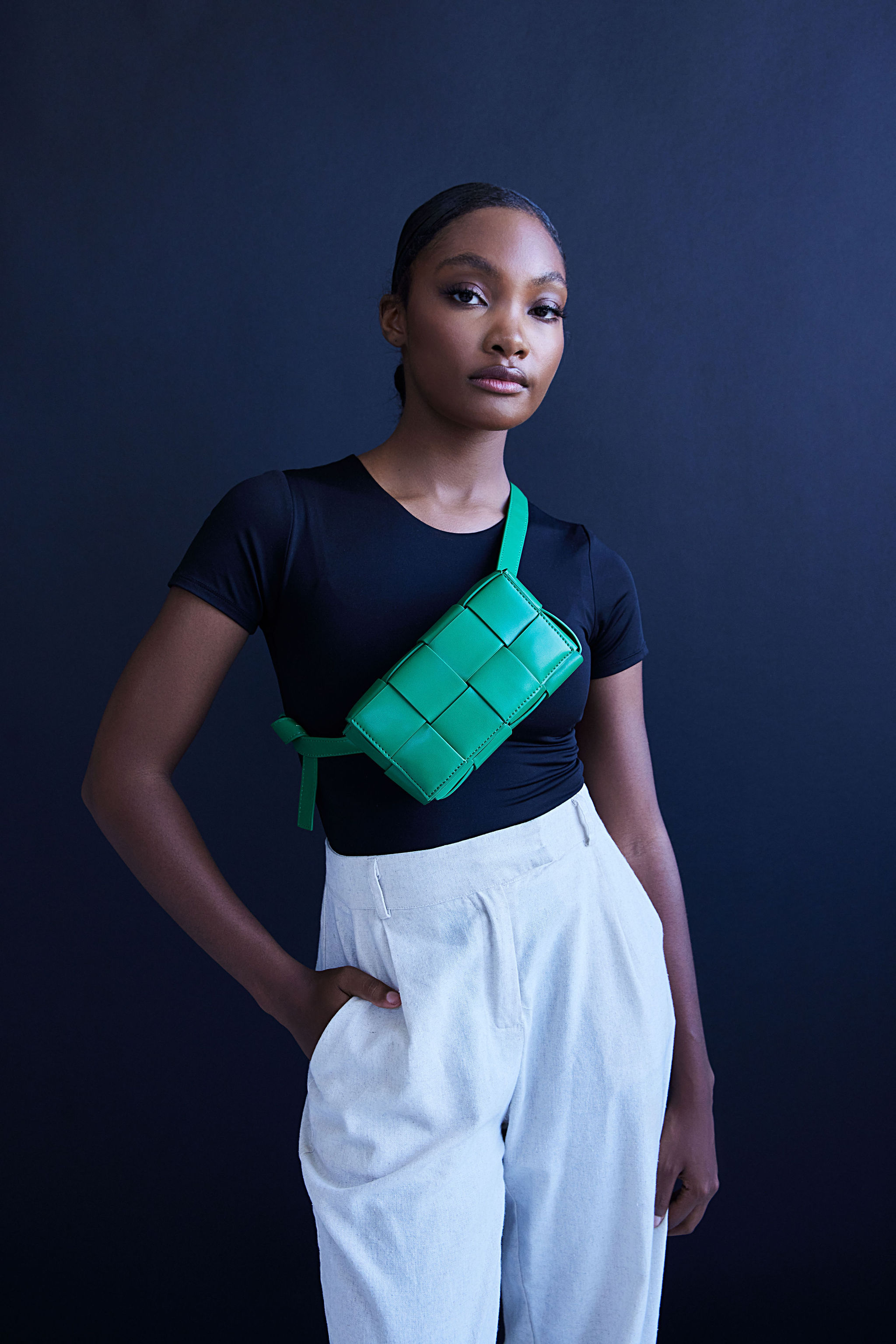 Portrait of a stylish woman in a black top and white pants wearing a green woven belt bag against a dark blue backdrop.