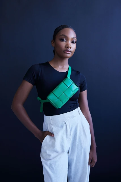 Portrait of a stylish woman in a black top and white pants wearing a green woven belt bag against a dark blue backdrop.