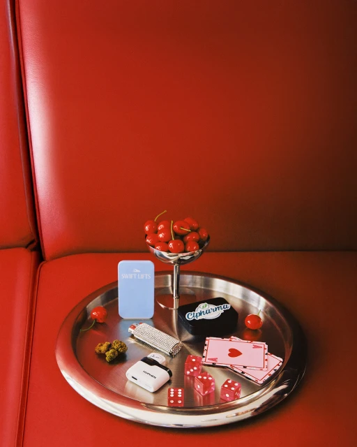 Close-up still life on a red surface featuring cherries in a silver bowl, dice, playing cards, and smoking accessories on a silver tray.