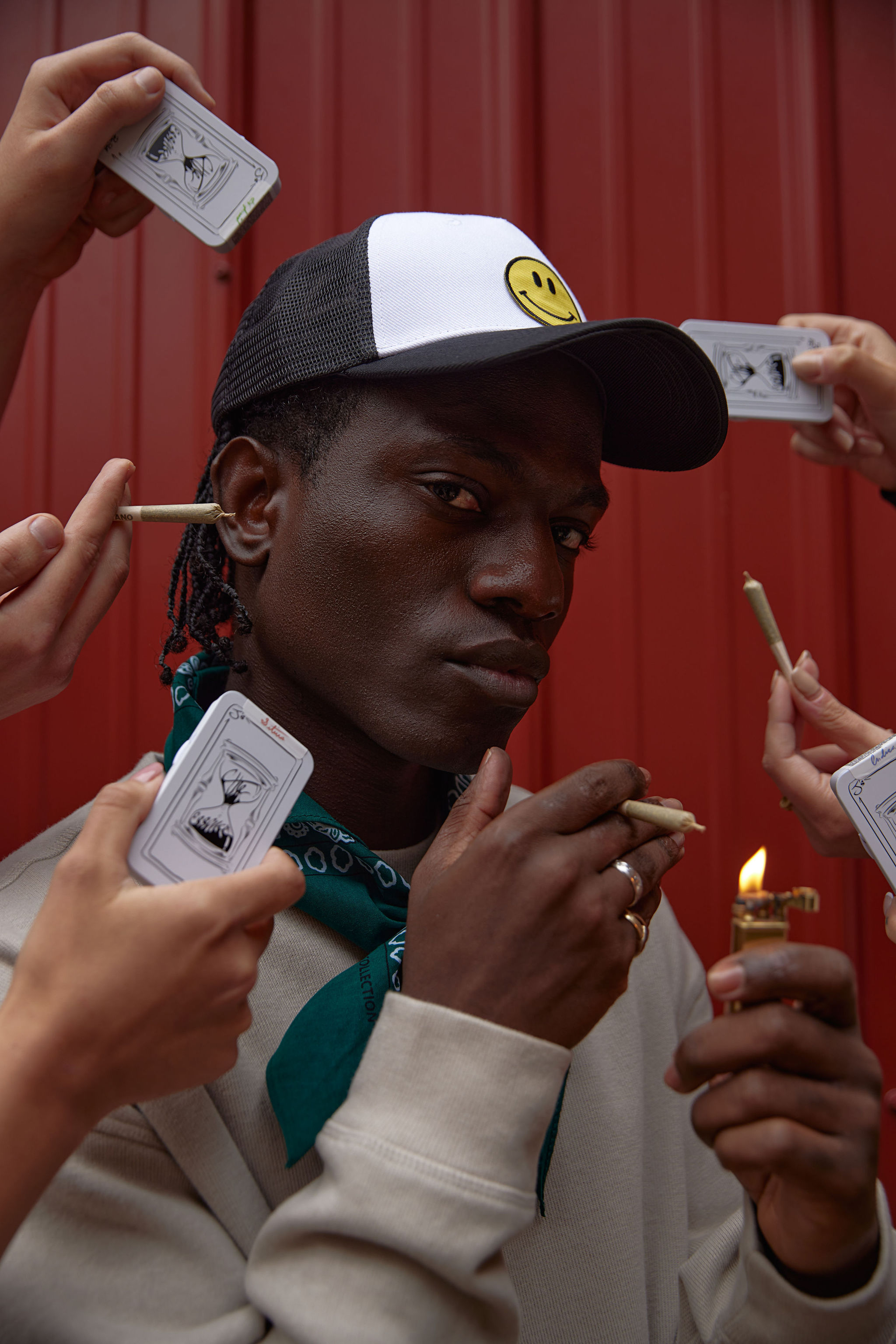A dark-skinned man with a white hat poses indoors surrounded by hands holding small tins and pre-rolled cigarettes, indoors.