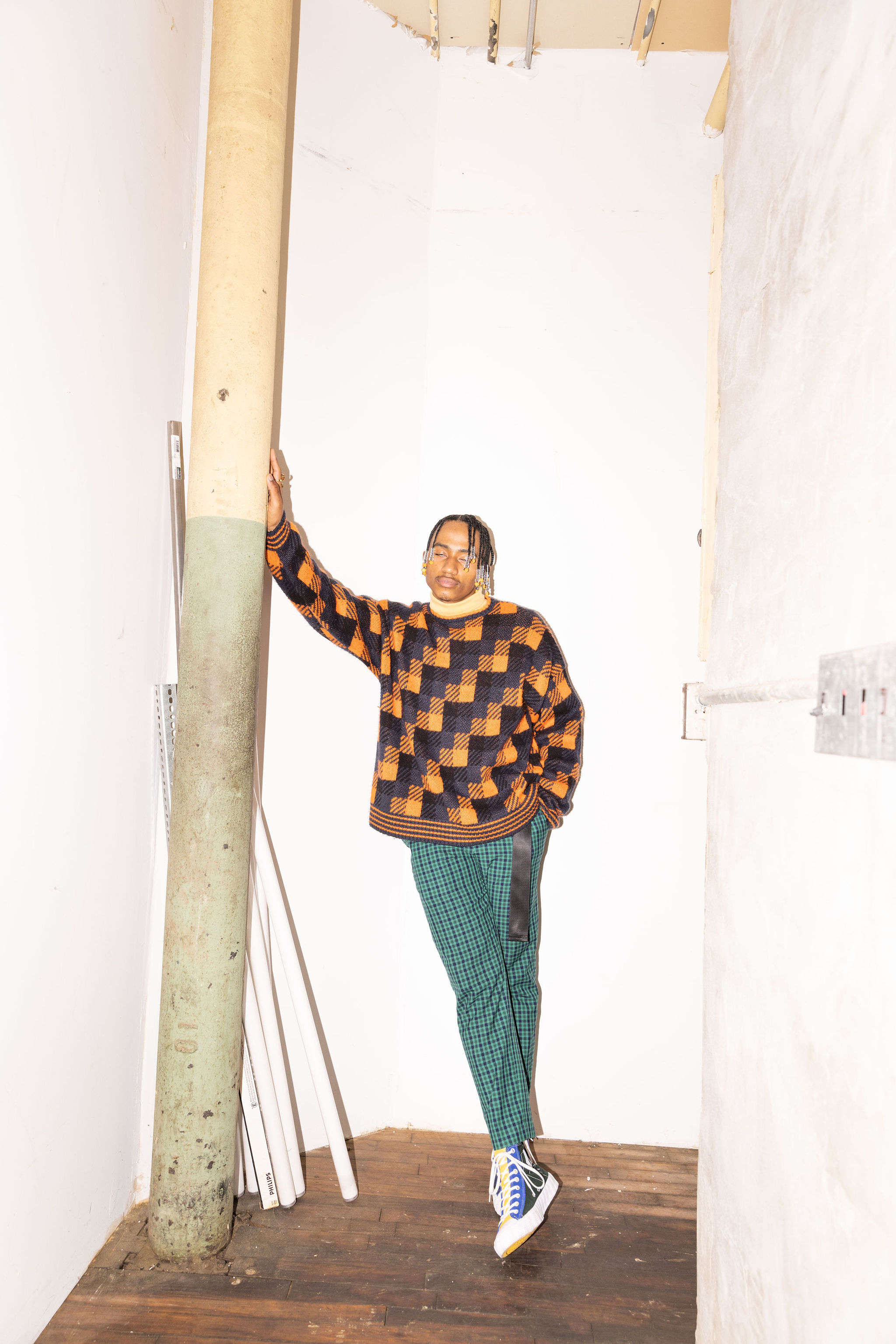 Young adult man poses indoors, leaning against a pipe in a checkered orange sweater, patterned pants, and sneakers in a casual fashion.