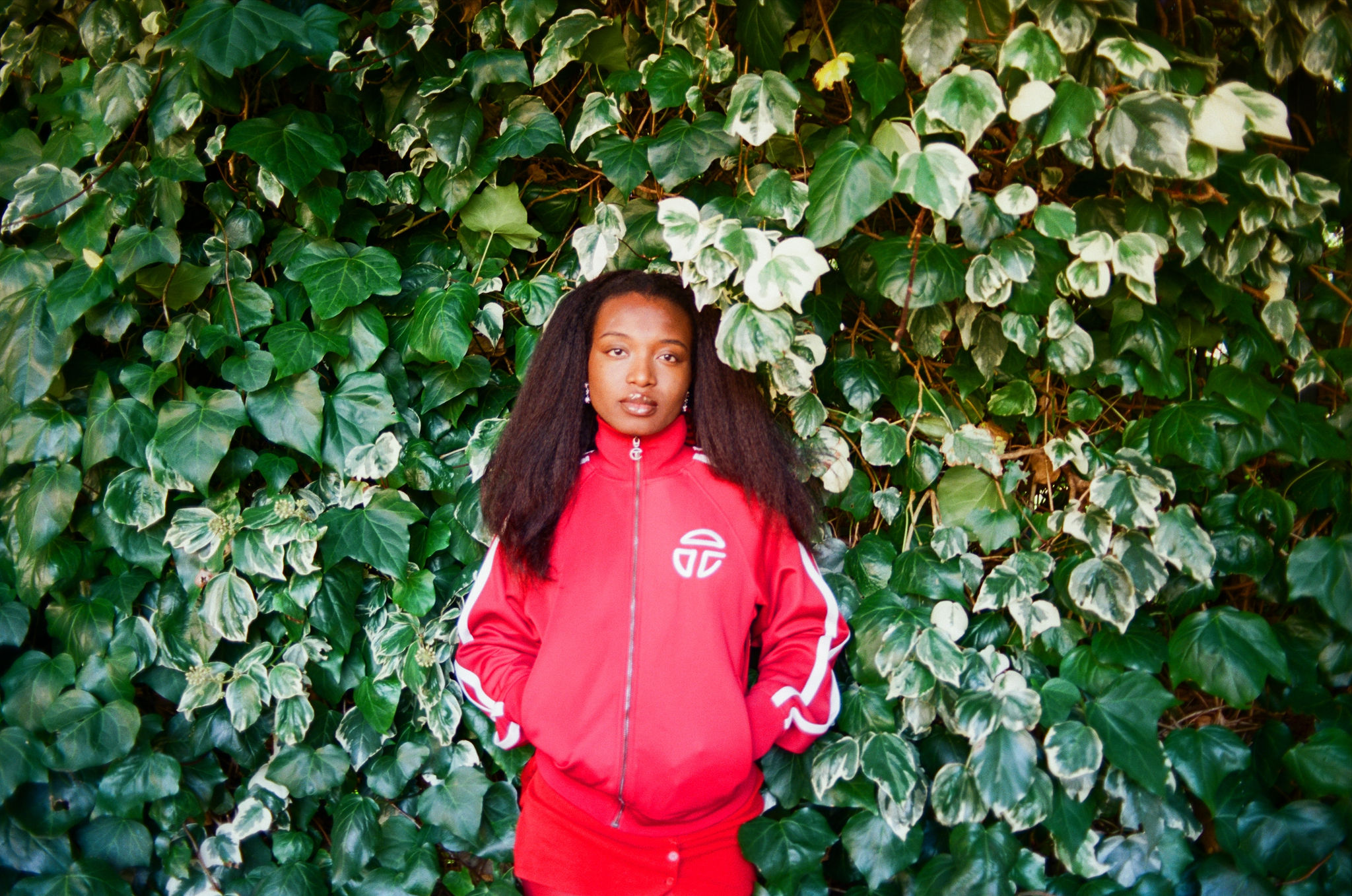 Portrait of a young Black woman in a red track suit against a lush green ivy wall, showcasing fashion and nature's backdrop.