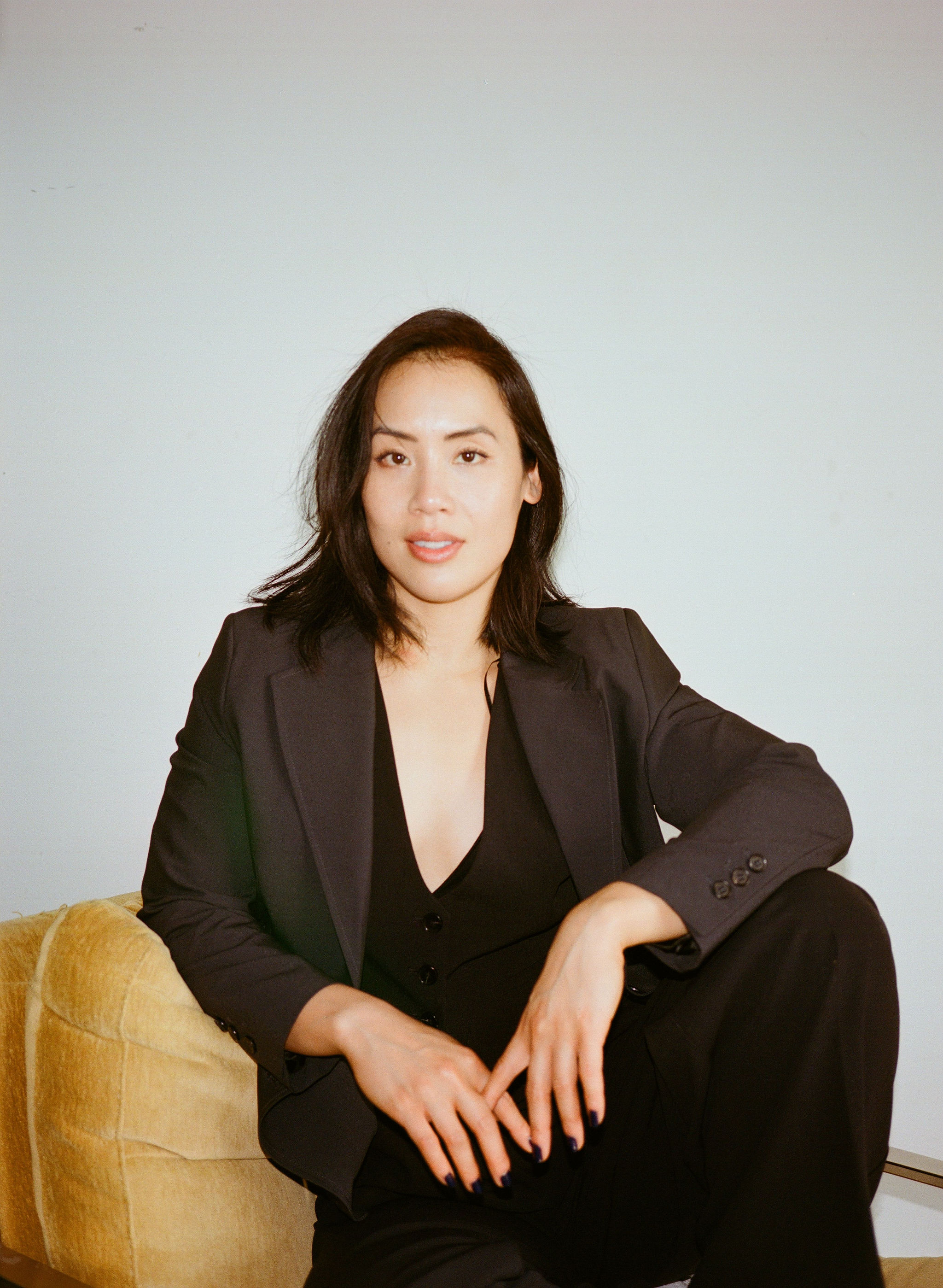 Portrait of a woman in a dark suit seated on a yellow chair against a pale blue background in a studio setting with soft lighting.