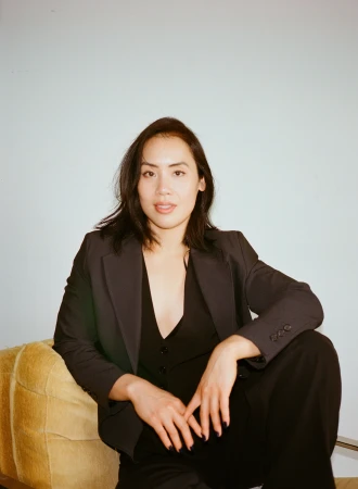 Portrait of a woman in a dark suit seated on a yellow chair against a pale blue background in a studio setting with soft lighting.