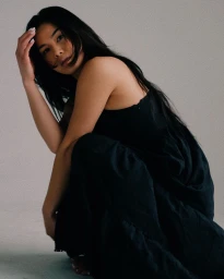 Woman with long dark hair wearing a black dress crouching against a plain grey backdrop in studio portrait