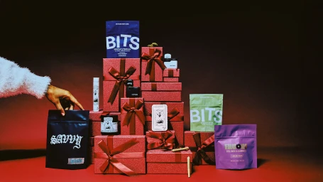A person's hand reaches for a dark pouch placed in front of stacked red gift boxes tied with ribbons and colorful product packages.