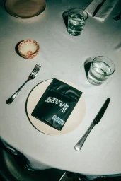 Overhead shot of a white-clothed dinner table featuring a black pouch with white gothic lettering sitting on a cream plate with cutlery and glassware