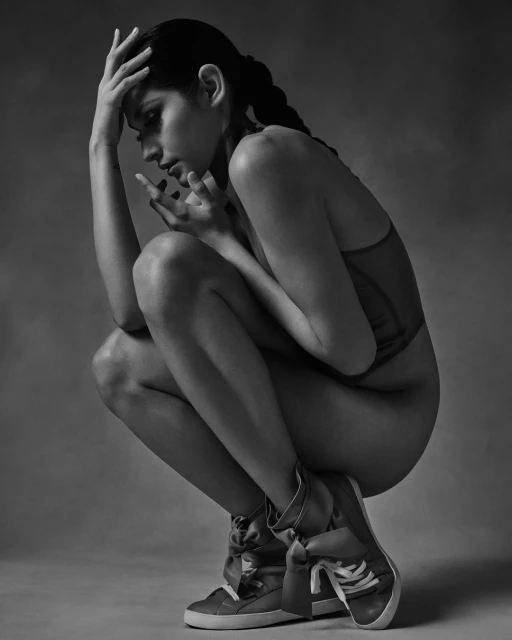 A young adult woman with braided hair crouches indoors wearing fashion sneakers and a bodysuit in a black and white, studio fashion portrait.