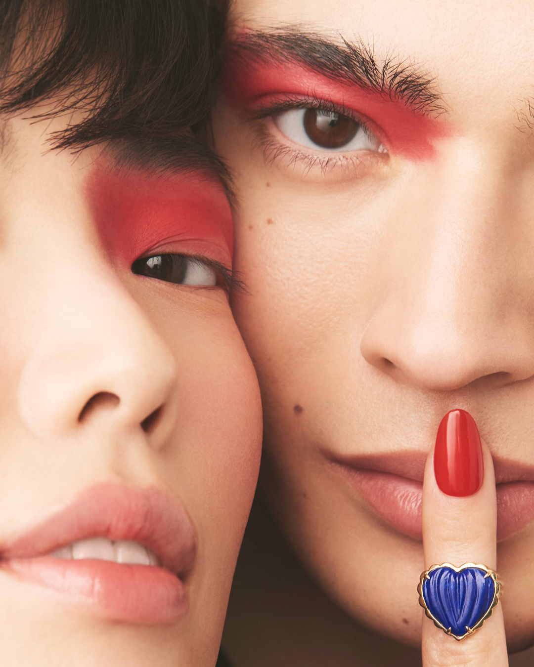 Close-up of two people with matching red eyeshadow and a red fingernail in the foreground wearing a blue heart ring.