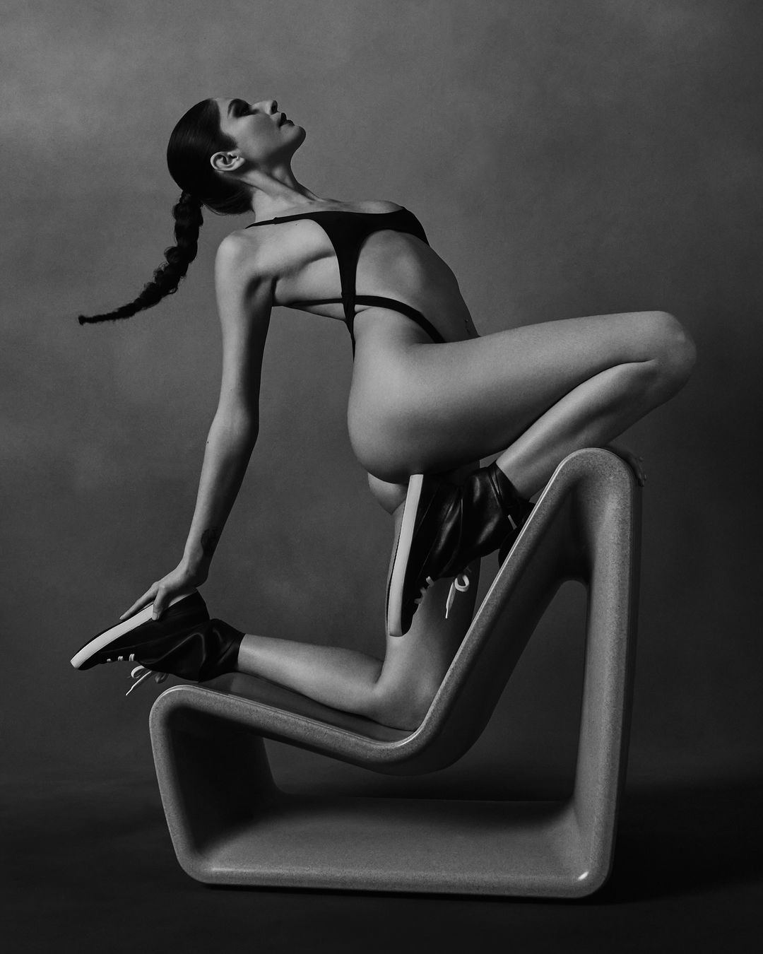 A woman in a black bodysuit and sneakers poses on a modern chair in a studio setting with a grey backdrop. She faces upward, with hair in a braid.