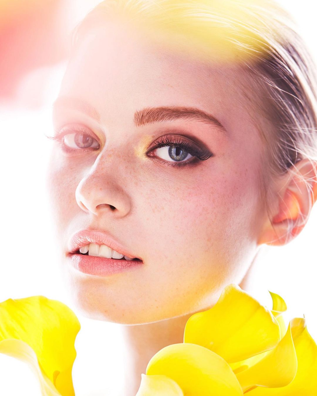 Close-up of a young woman's face with fair skin, freckles, and light hair, accented by yellow floral petals around her neck and shoulders.