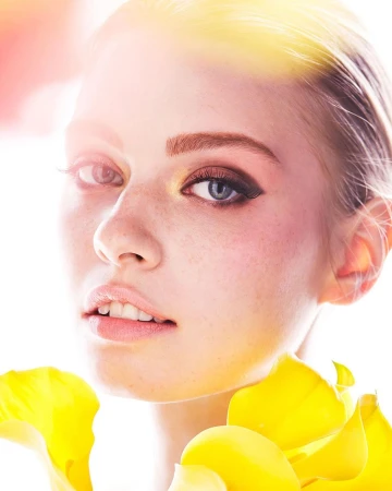 Close-up of a young woman's face with fair skin, freckles, and light hair, accented by yellow floral petals around her neck and shoulders.