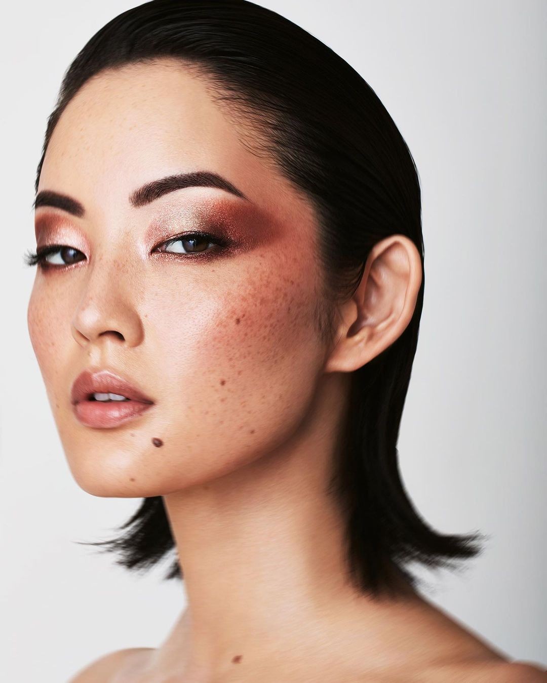 Close-up portrait of an Asian woman with slicked-back dark hair, warm eyeshadow, and fair skin against a bright, neutral background.