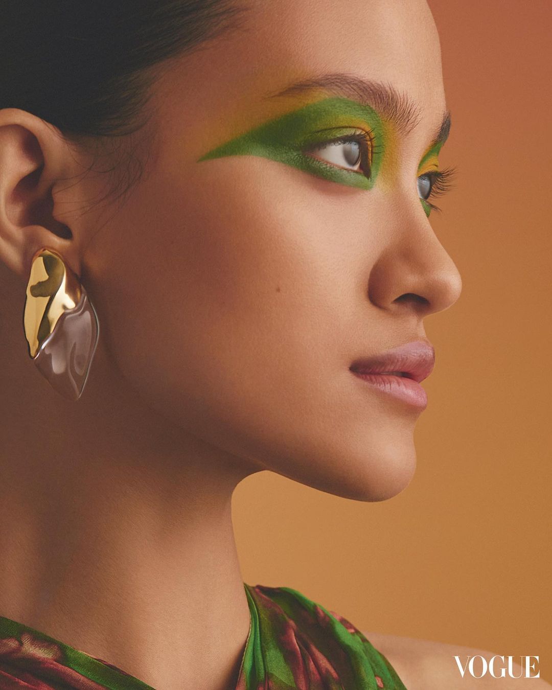 Close-up profile of a woman with green eyeshadow and gold earrings against an orange background for beauty and fashion.