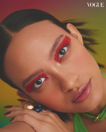 Close-up portrait of a woman with bold red eye makeup and green clothing against a gradient background.