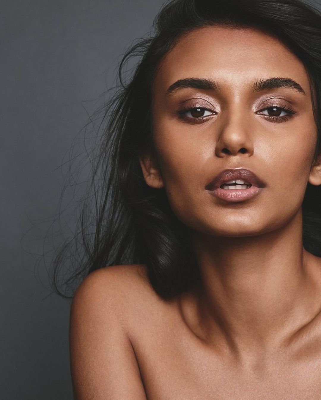 Close-up portrait of a stunning South Asian woman with dark hair and glowing skin, captured against a neutral gray backdrop in a beauty context.