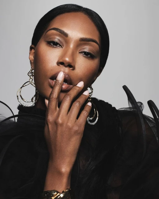Close-up portrait of a woman with dark hair and brown skin in a black sheer top, gold earrings, and a gold bracelet against a grey backdrop.