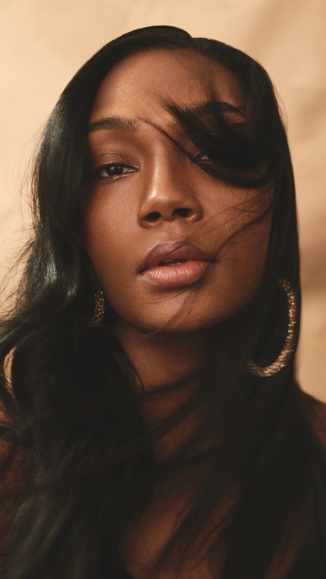 Close-up portrait of a woman with long dark hair blowing across her face and gold hoop earrings against a warm textured background