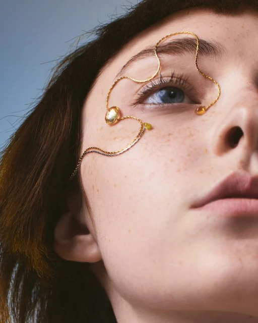Portrait of a woman's face with a delicate gold chain accenting her eyebrow and eye, set against a soft light blue backdrop.
