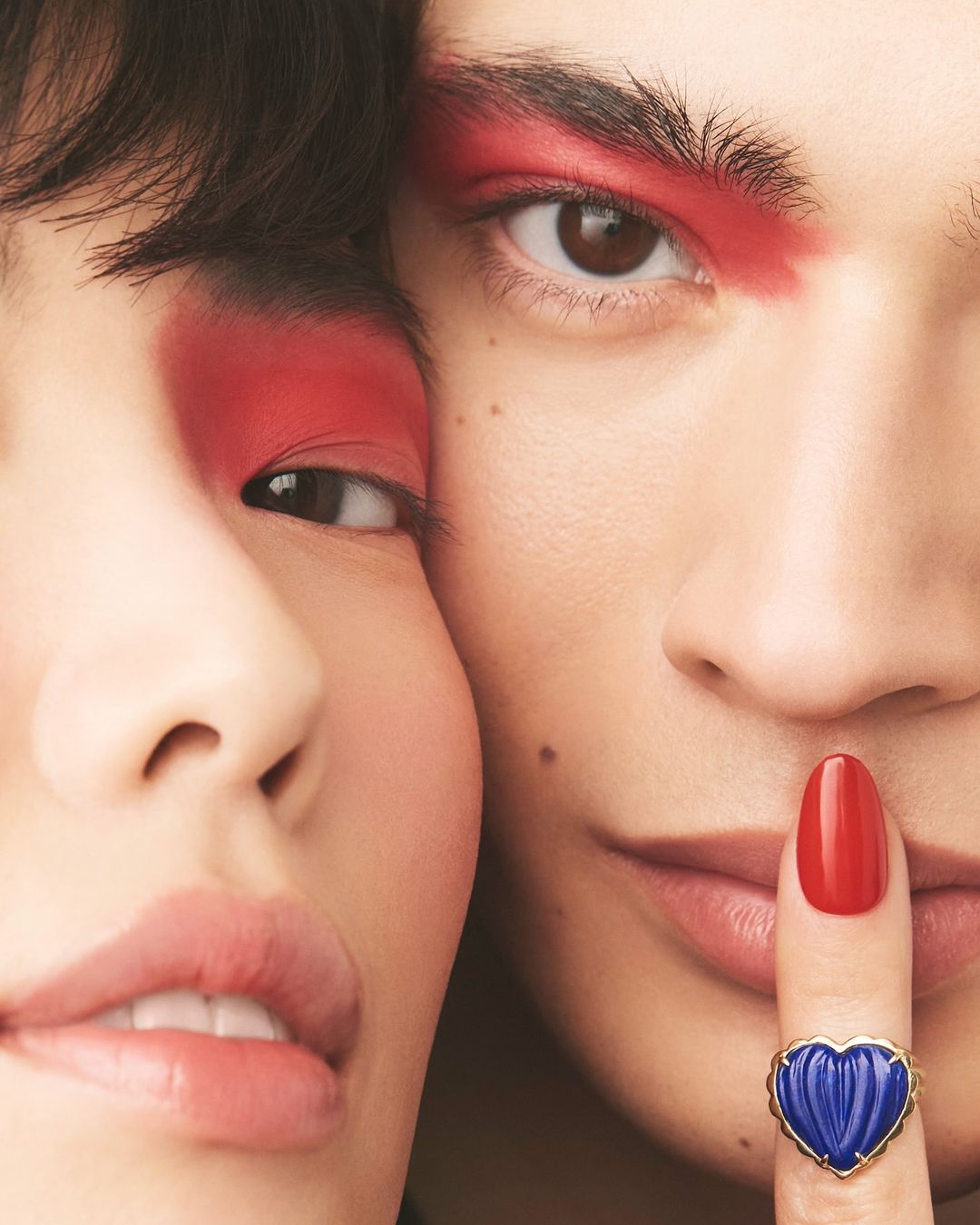 Close-up of two faces with red eye makeup and a hand with red nails and blue heart ring up to one person's lips.