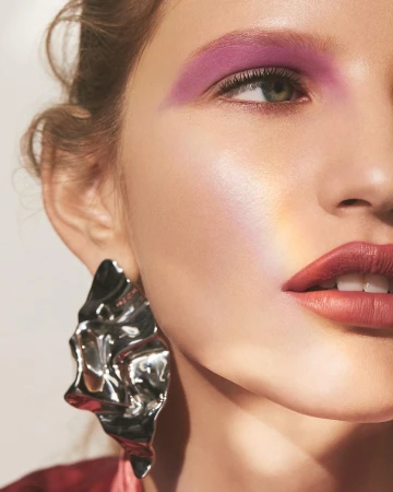 Close-up portrait of an adult female model with violet eyeshadow, dark pink lipstick, and a statement silver earring.
