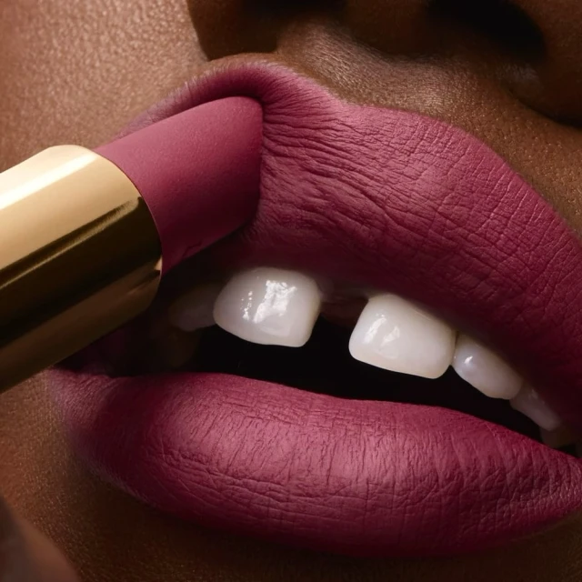 Close-up of lipstick being applied to dark-skinned female model's lips; vibrant rose-pink matte lipstick, gold tube, flawless skin, beauty shot.