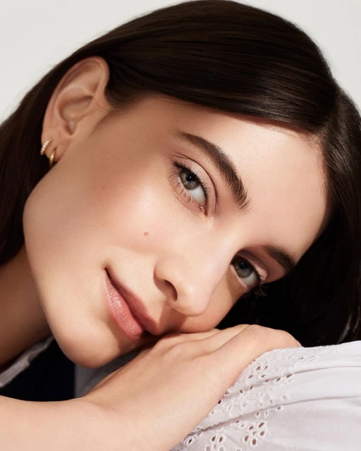 Close-up portrait of a dark-haired woman with light skin and a mole, resting her cheek on a white, embroidered fabric surface.