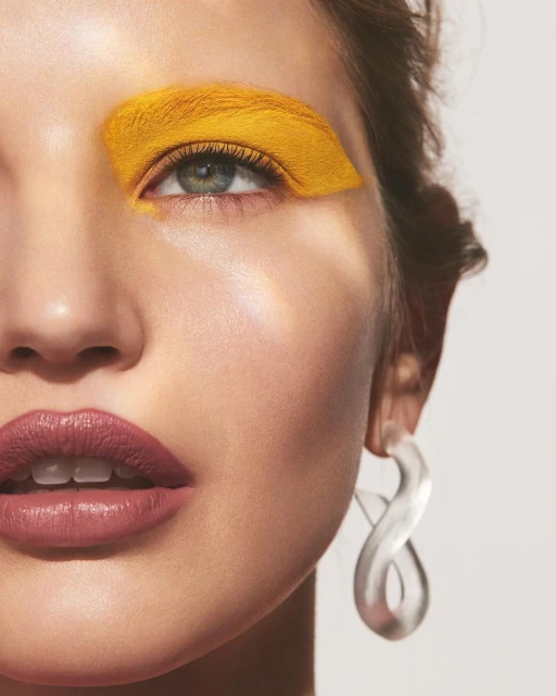Close-up of a woman's face with yellow eyeshadow and neutral lipstick, showing her clear earring in front of a white background.
