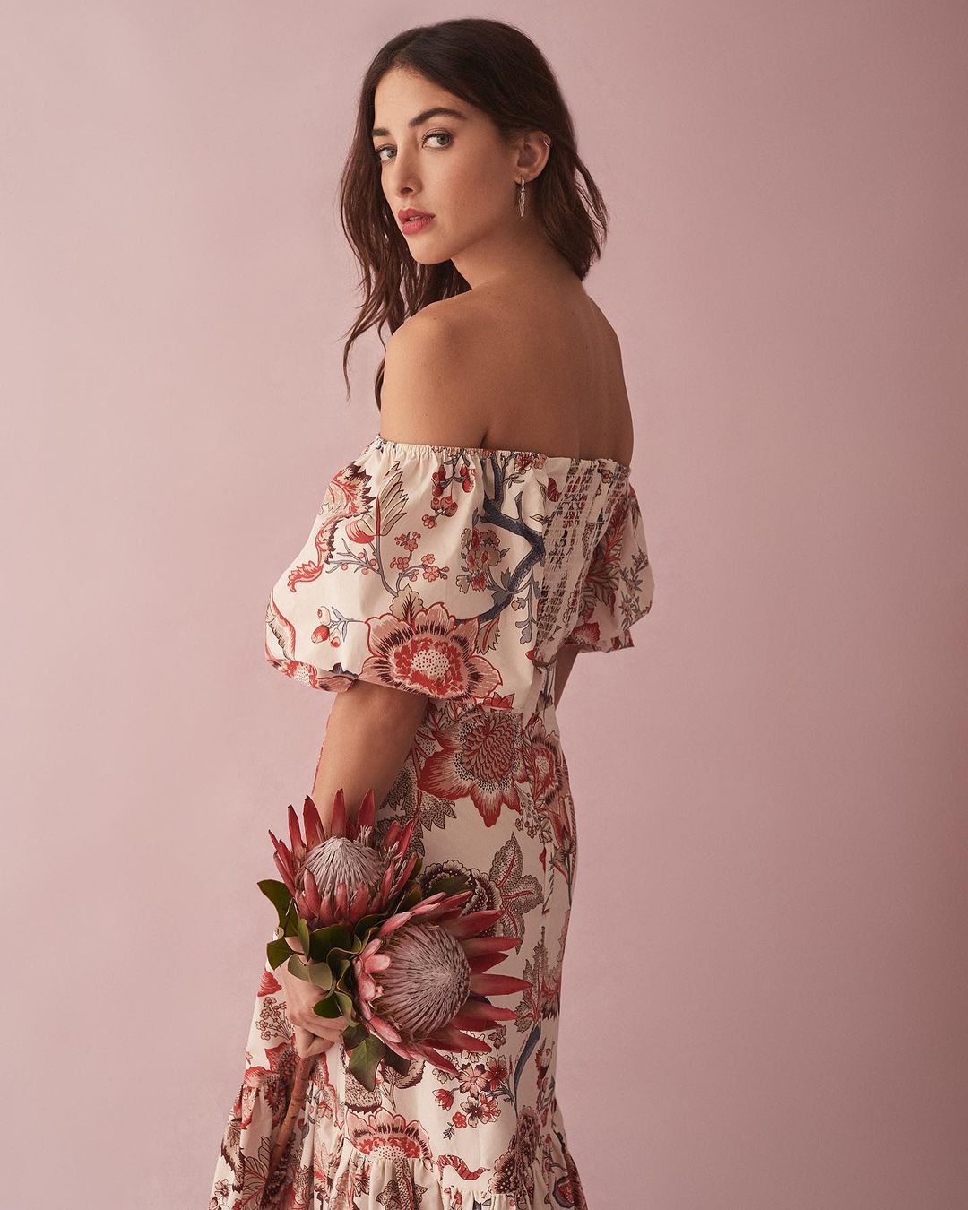Elegant woman in floral dress with off-the-shoulder neckline holding two red protea flowers against soft pink backdrop in studio shot.