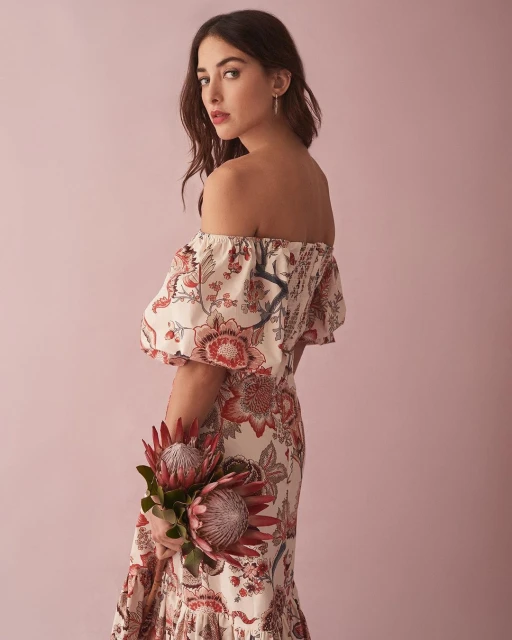 Elegant woman in floral dress with off-the-shoulder neckline holding two red protea flowers against soft pink backdrop in studio shot.