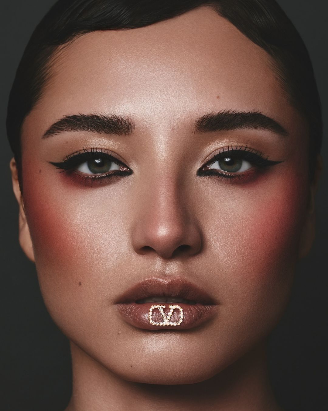 Close-up portrait of a woman with dark hair and dramatic eye makeup, featuring a glittering V logo lip accessory against a dark background.