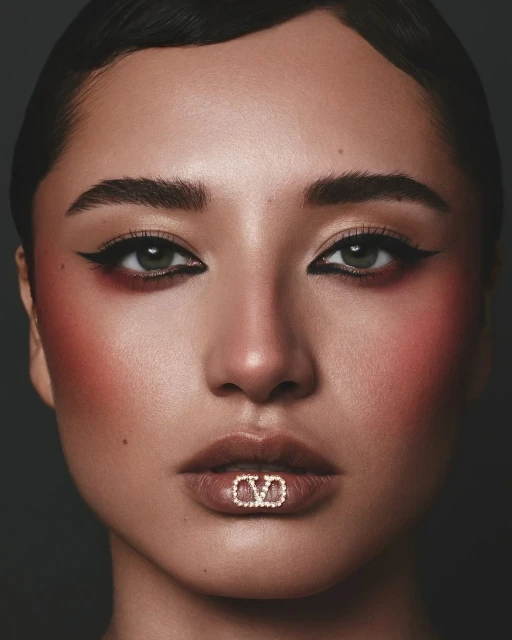 Close-up portrait of a woman with dark hair and dramatic eye makeup, featuring a glittering V logo lip accessory against a dark background.