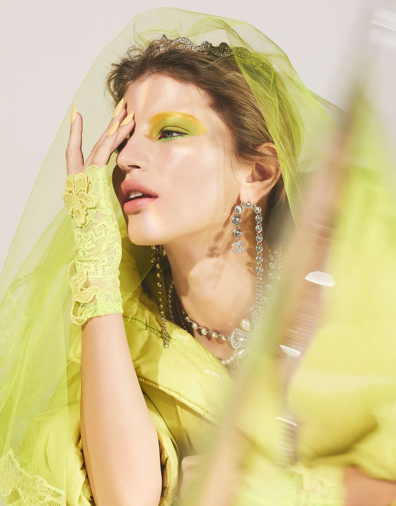 Close-up portrait of woman wearing a yellow veil, bright yellow eye makeup, pearl jewelry, and lace gloves in a studio.