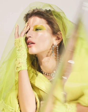 Close-up portrait of woman wearing a yellow veil, bright yellow eye makeup, pearl jewelry, and lace gloves in a studio.