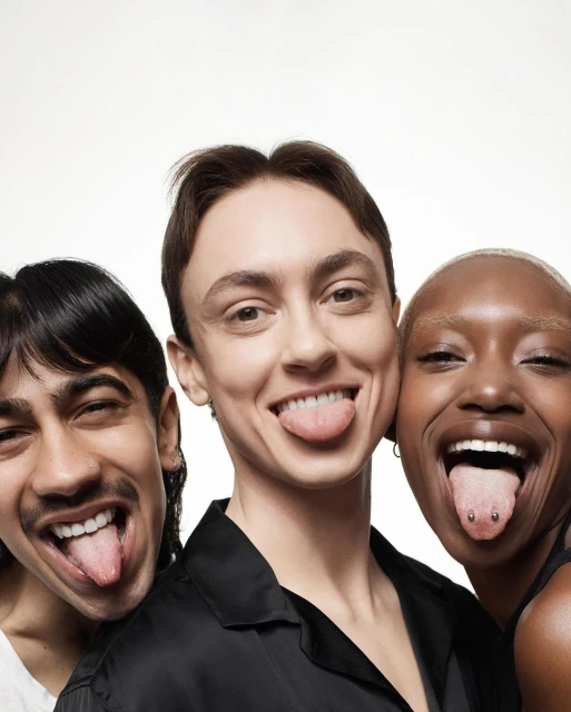 Three smiling people posing closely together, sticking out their tongues in a light photo studio setting. Adult male and female are visible.
