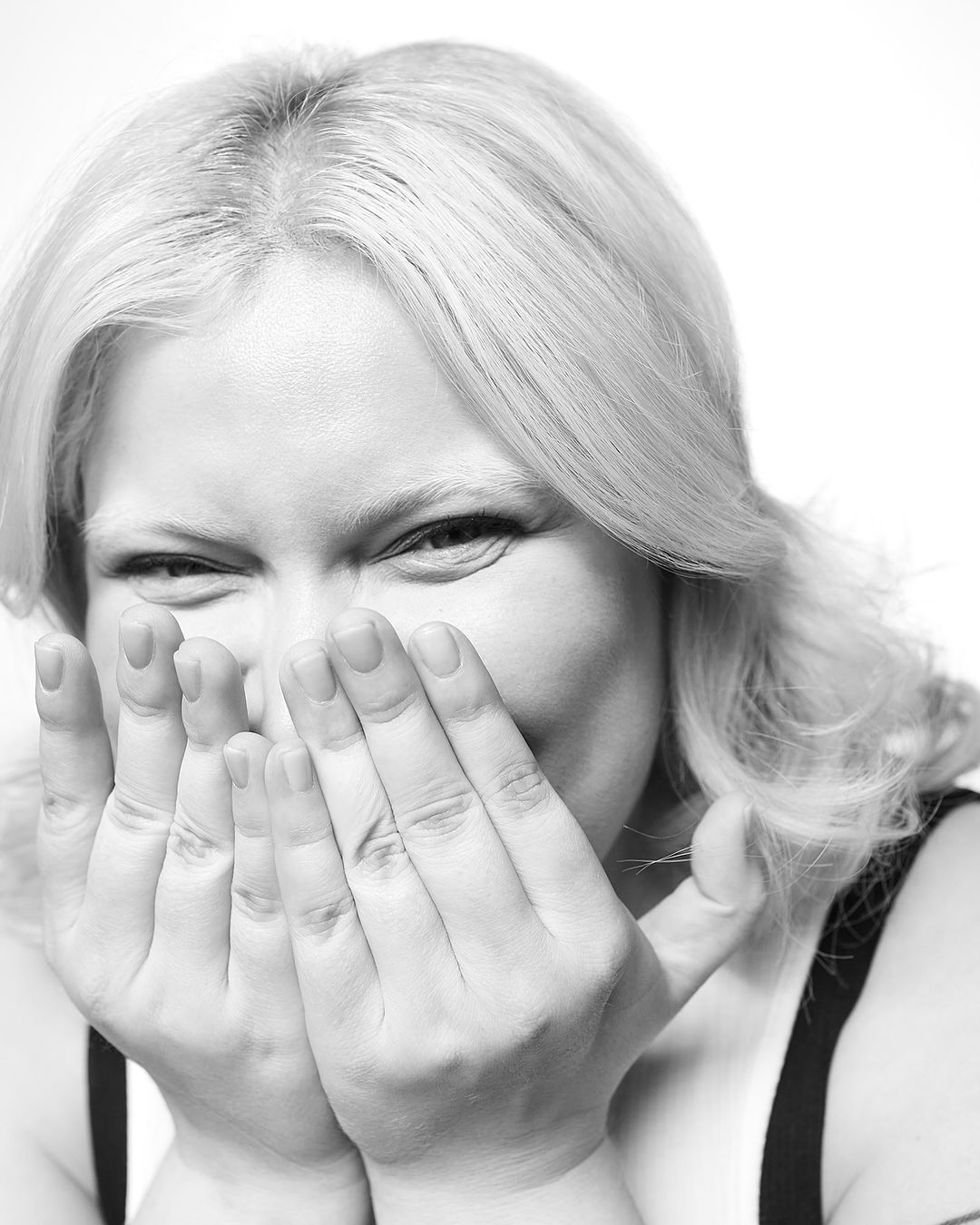 Monochrome portrait of a blonde adult female with hands covering her mouth, set against a bright white background in a photo studio.