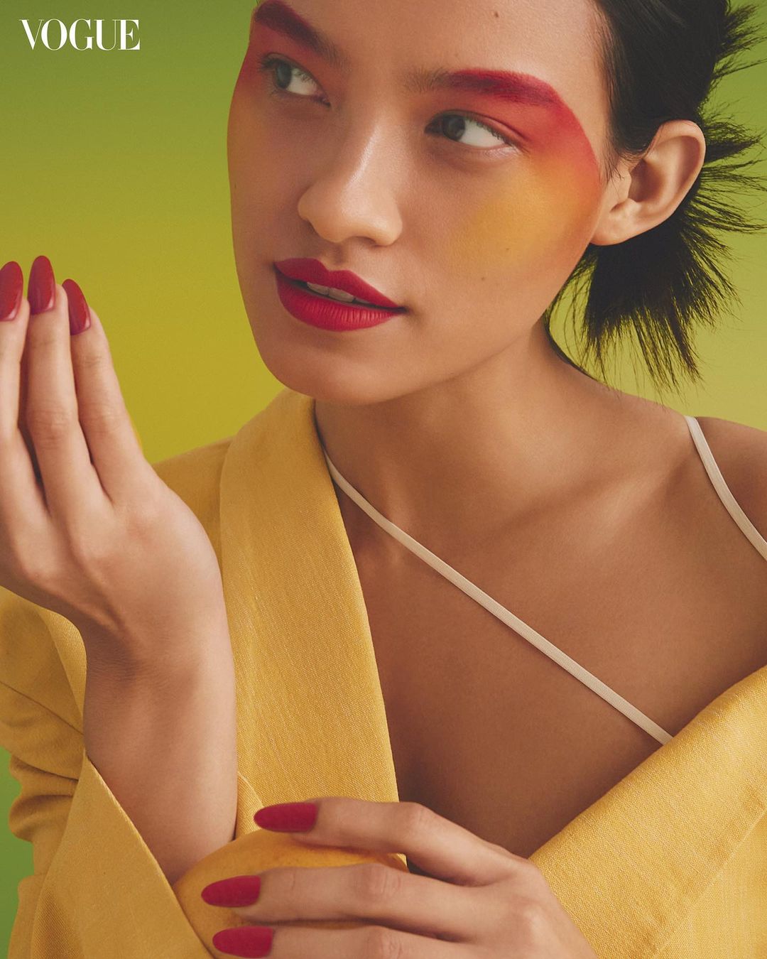 Close-up of a stylish Asian woman with vibrant makeup and red nails, wearing a yellow blazer against a lime green background.