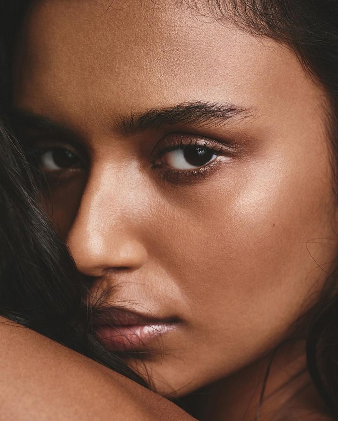 Close-up portrait of a young adult woman with brown hair, brown eyes, and brown skin, photographed in soft studio lighting.