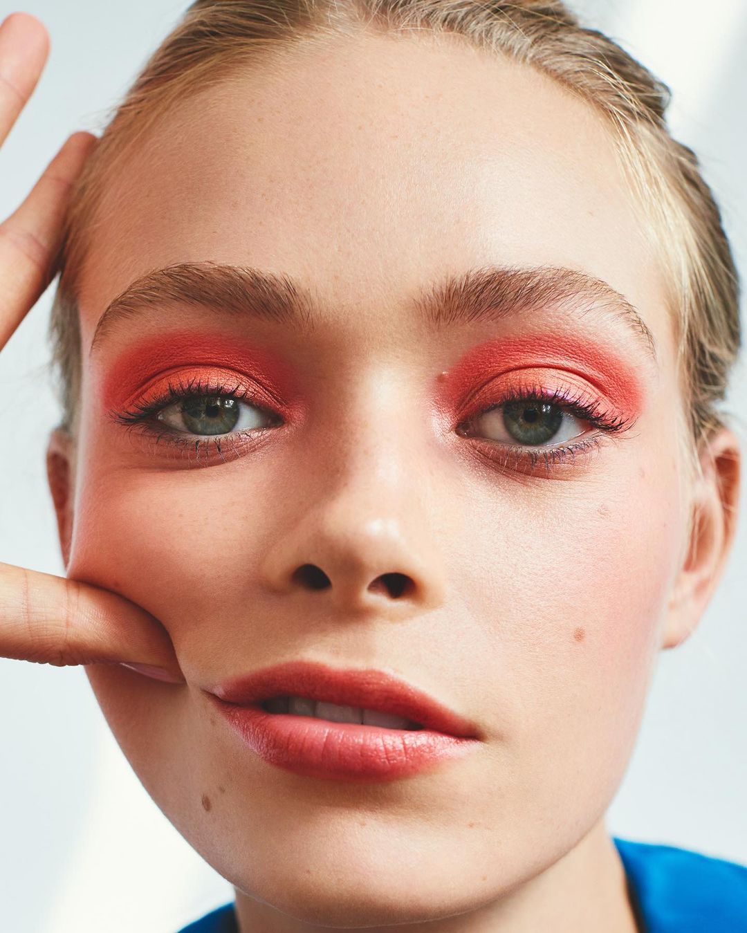 Close-up portrait of a young woman with red eyeshadow and fair skin. An adult hand touches her face in a beauty shot.