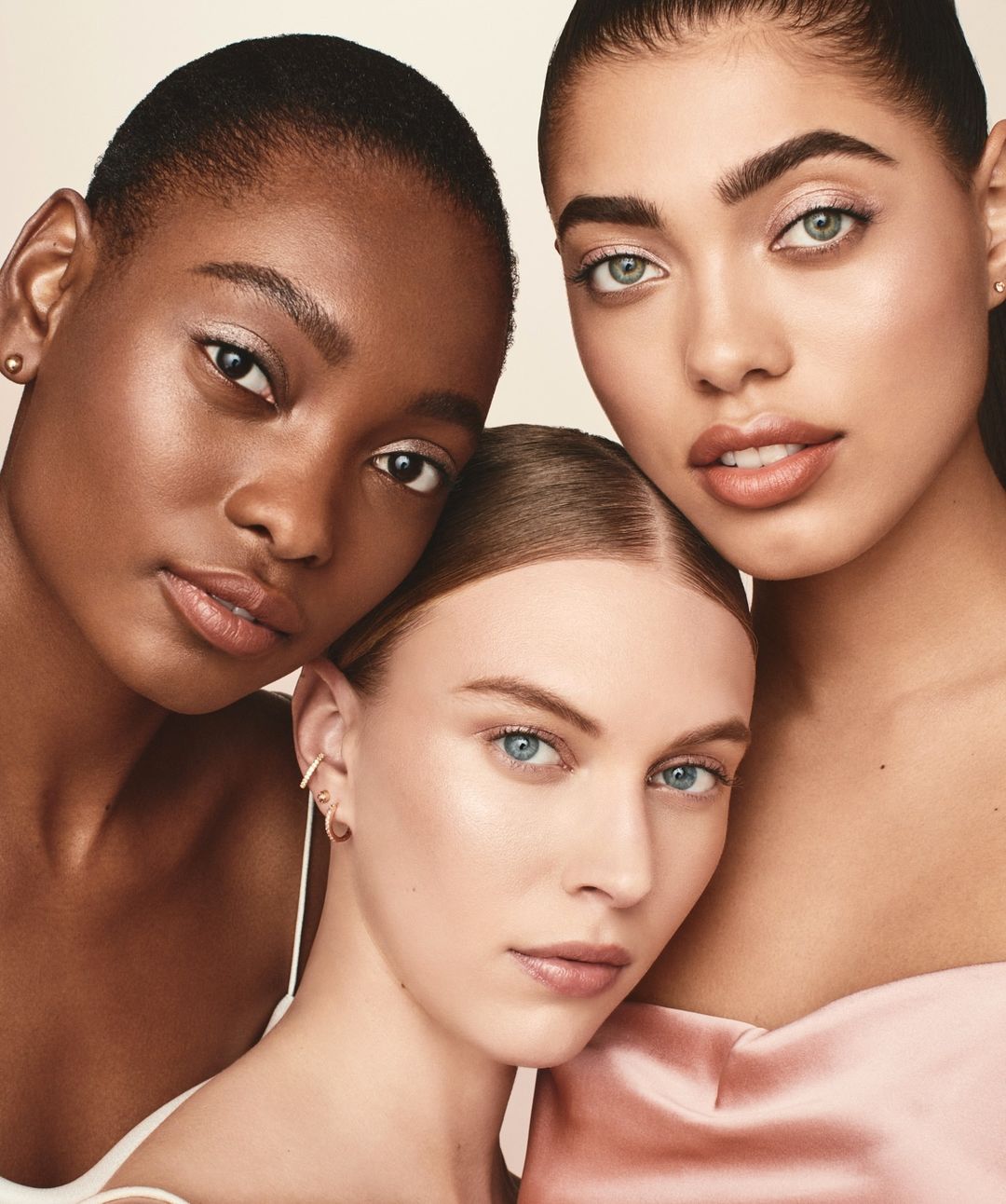 Close-up studio shot of three attractive young adult women with diverse skin tones, wearing fashionable makeup and jewelry, posing closely together.