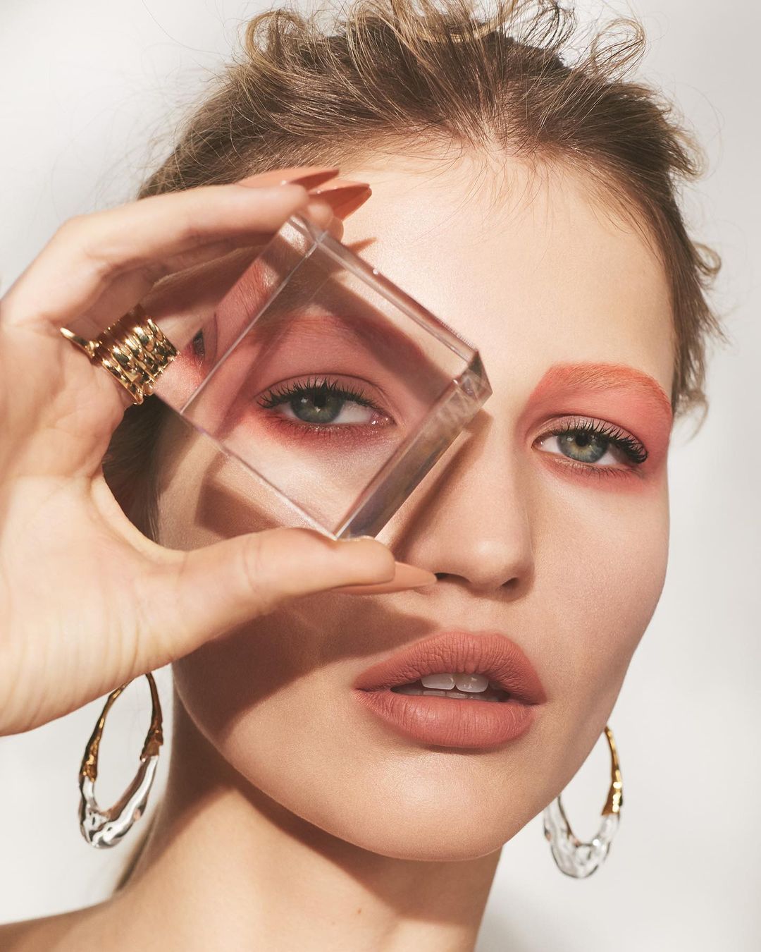 Close-up portrait of a woman with geometric makeup, holding a clear cube in front of her eye, and wearing large clear earrings.