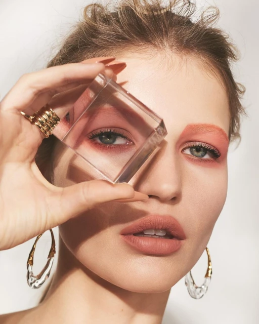 Close-up portrait of a woman with geometric makeup, holding a clear cube in front of her eye, and wearing large clear earrings.