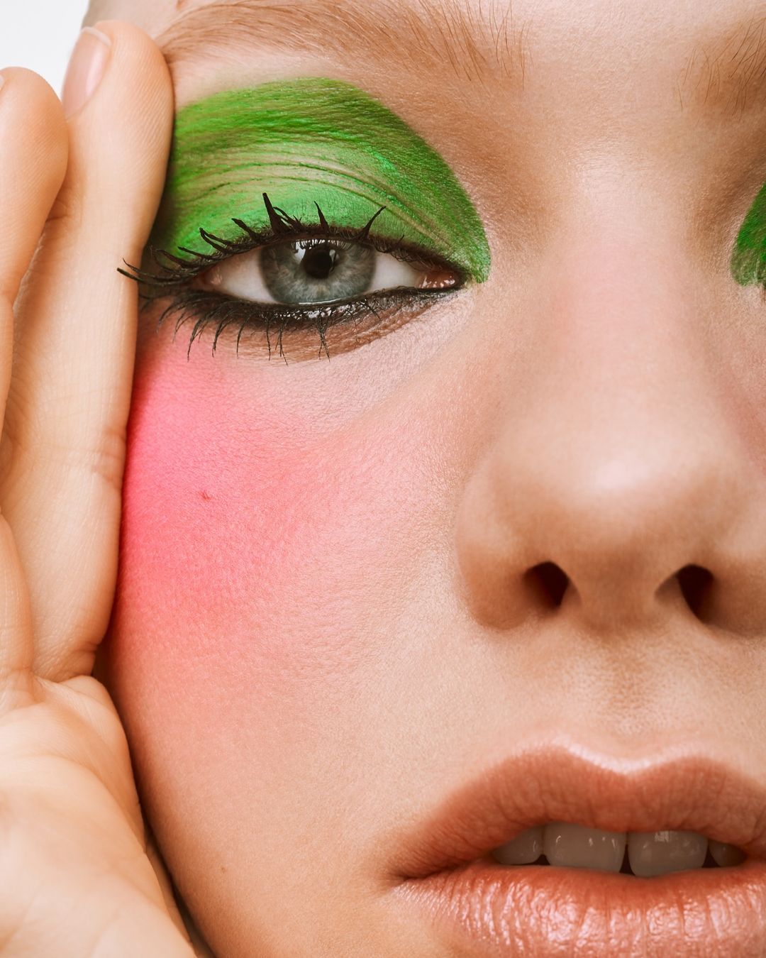 Close-up of a woman's face with striking green eye shadow and bright pink blush, showcasing bold and colorful makeup.