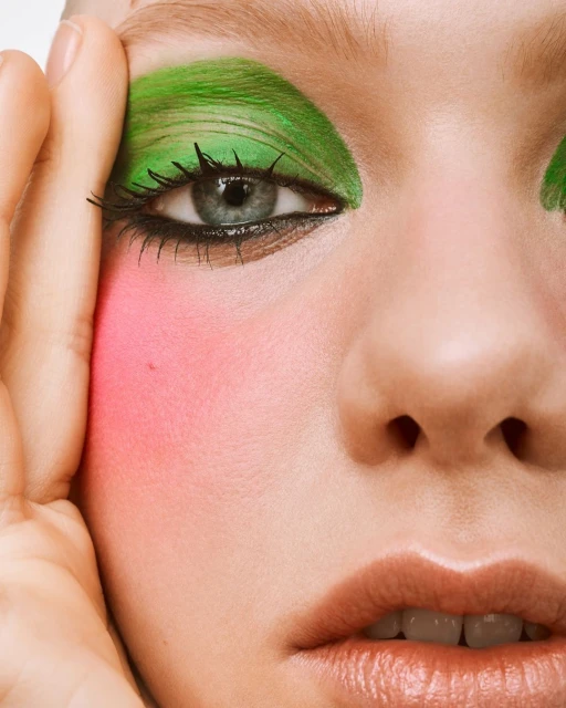 Close-up of a woman's face with striking green eye shadow and bright pink blush, showcasing bold and colorful makeup.